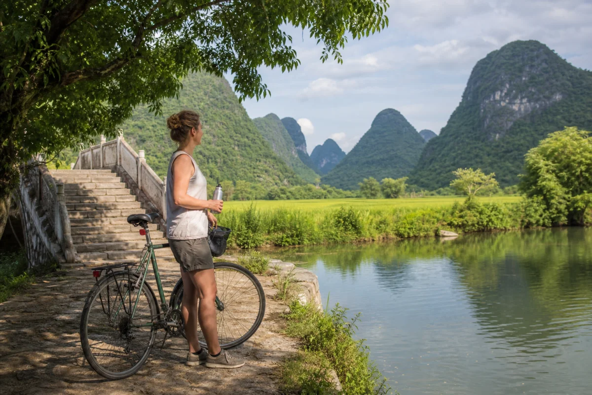 Traveler pausing beside a Yangshuo countryside river with a bicycle